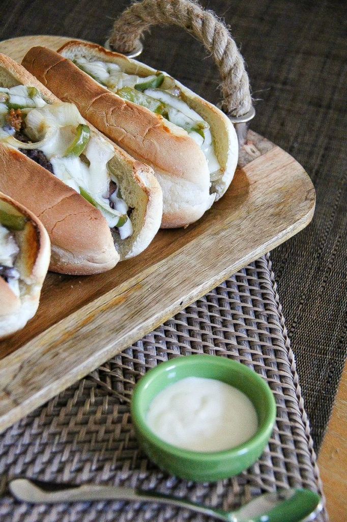 philly cheesesteak sandwiches on a wood serving tray with a bowl of horseradish sauce on the side