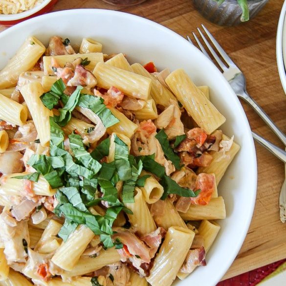 rigatoni pasta with chicken and bacon surrounded by red pepper flakes, grated cheese, and fresh herbs