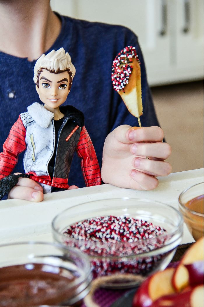 hand holding an apple slice on a stick dipped in chocolate and sprinkles with a carlos descendants doll