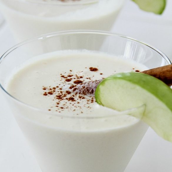 creamy cocktail in a glass with an apple slice and cinnamon stick garnish