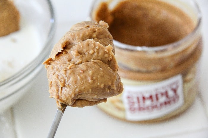 a spoon of peanut butter with a jar of simply ground peanut butter in the background