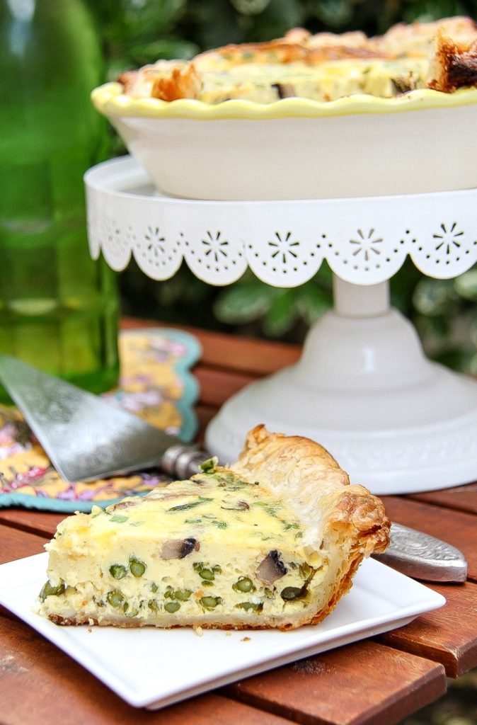 A cake stand with an asparagus and mushroom quiche with a slice on a plate underneath it