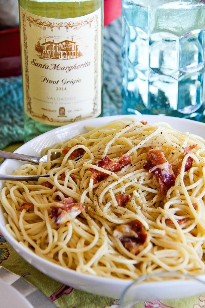 spaghetti carbonara and bacon in a white bowl with a bottle of pinot grigio in the background