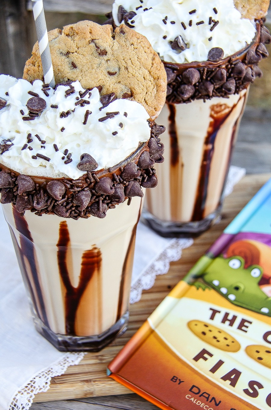 An over the top chocolate chip cookie milkshake topped with whipped cream and a chocolate chip cookies with a glass rimmed with chocolate chips and sprinkles.
