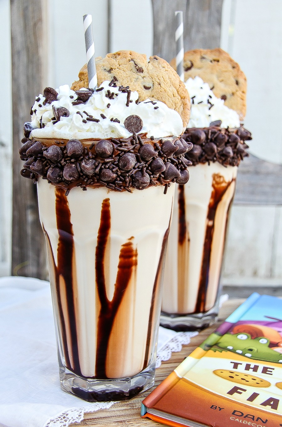 a chocolate chip cookie milkshake with chocolate drizzled inside the glass, the glass rimmed with chocolate chips and the milkshake topped with whipped cream and a chocolate chip cookie