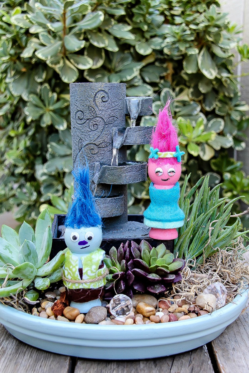 Handmade Trolls inside a container fairy garden with succulents and a water feature. 