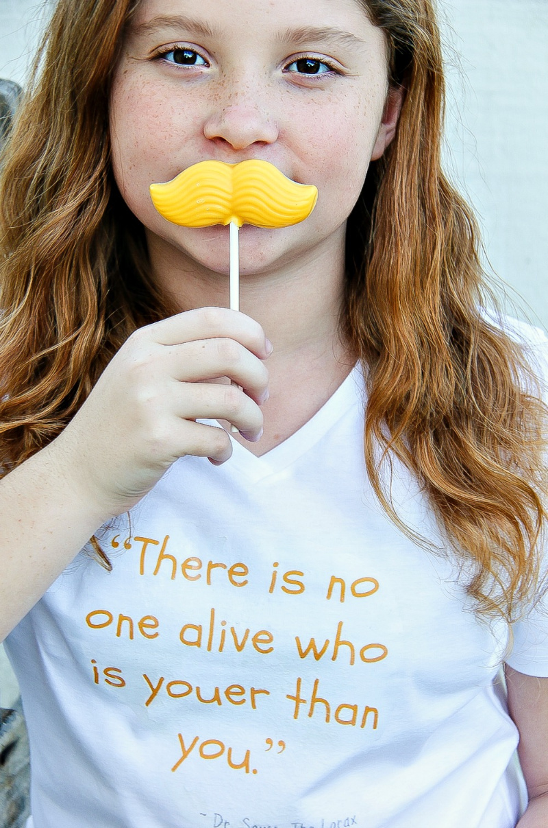 A girl wearing a tee with the lorax quote there is no one alive who is youer than you and holding a yellow mustache lollipop