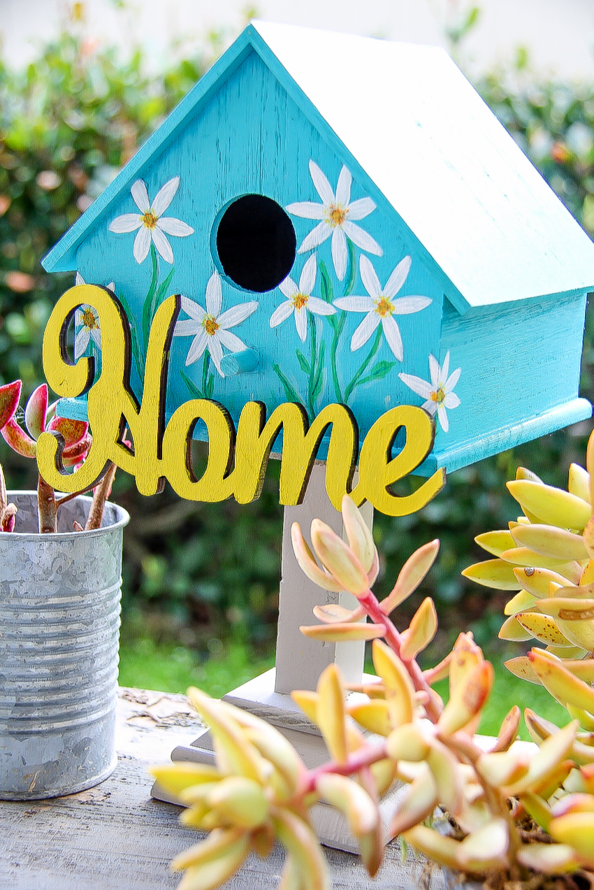 a birdhouse painted blue with flowers on it and a home sign in yellow