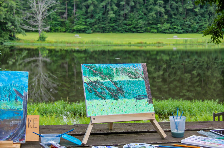 outdoor landscape painting on canvas is done by a lake.