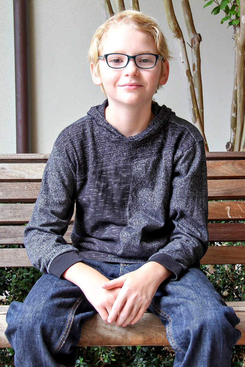 a boy wearing glasses sitting down wearing a grey hooded top and blue jeans