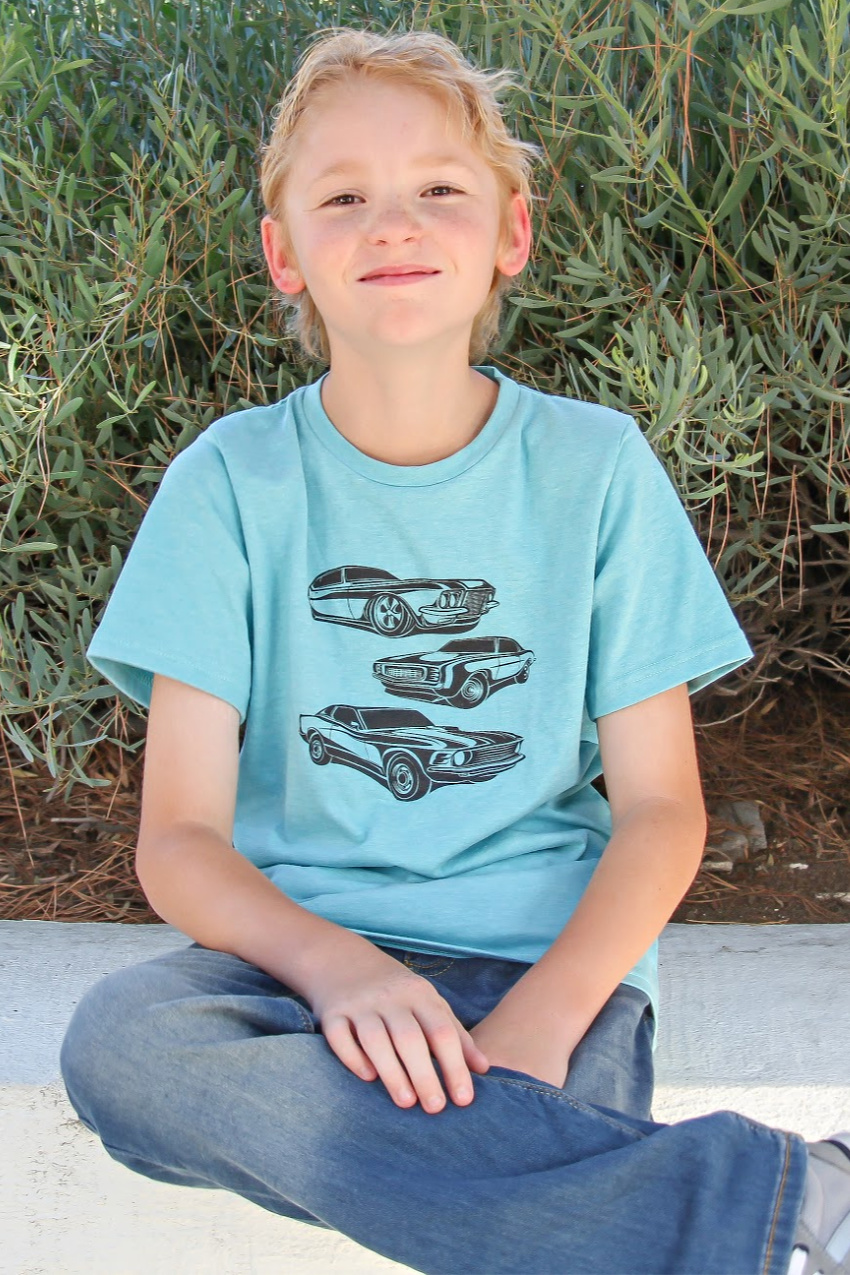 a boy sitting down wearing jeans and a light blue tee with cars on it