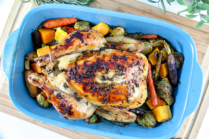 a whole roast chicken in a blue baking dish surrounded by roasted vegetables