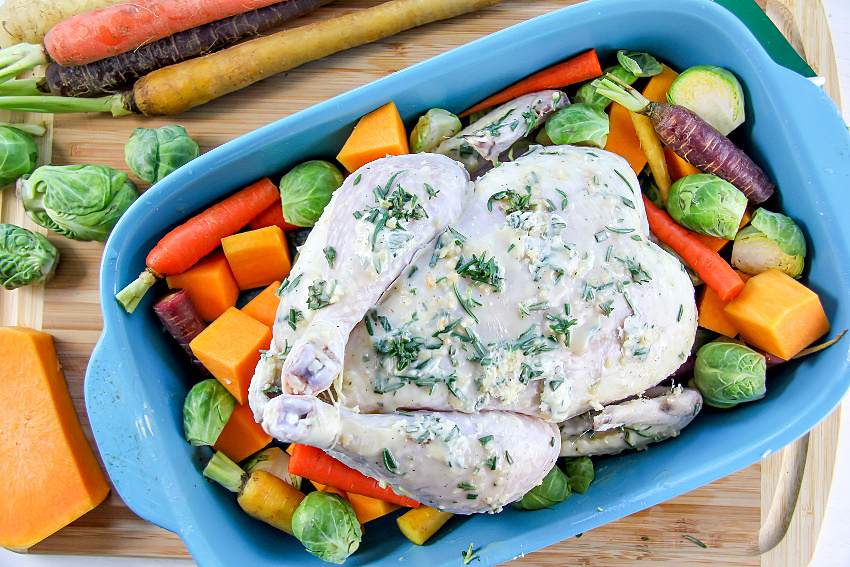 A whole chicken in a blue baking dish surrounded by fresh vegetables