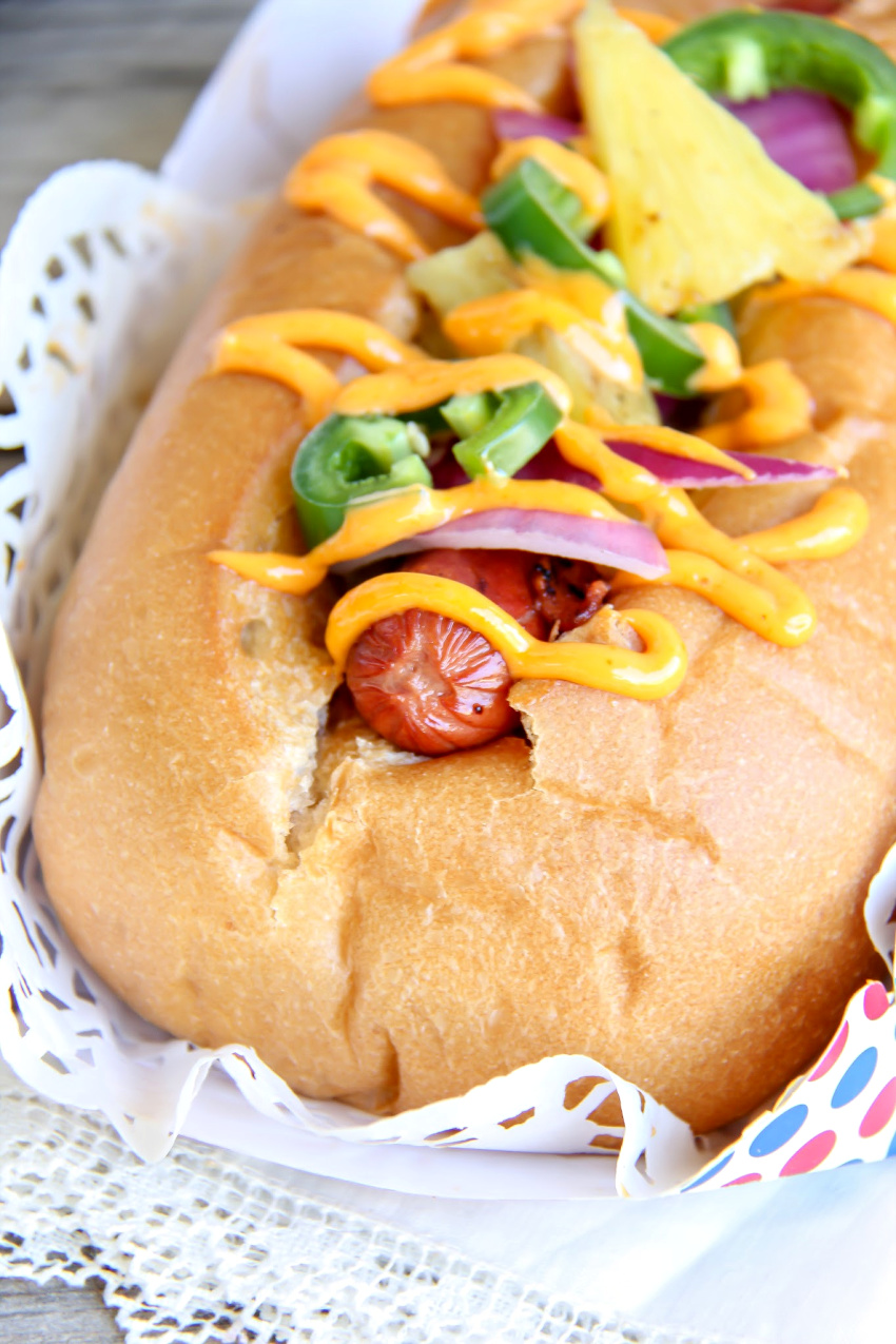 a hot dog in a bun topped with jalapeno, red onion, pineapple, and sriracha sauce