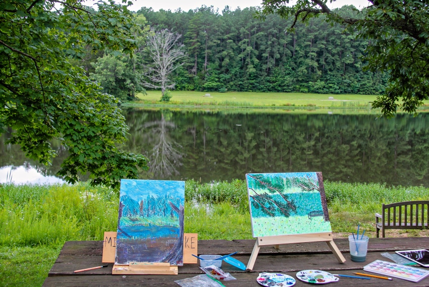 Daytime outdoor date idea. Painting landscapes with art easels and paints in front of a lake with green trees.