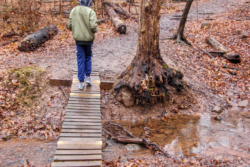 Outdoor winter activities in New Jersey with kids. A boy walking on a plank bridge over a stream with a small fairy house in a tree stump by the water