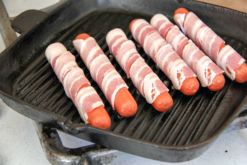 hot dogs that have been wrapped in bacon on an indoor grill pan on the stove.