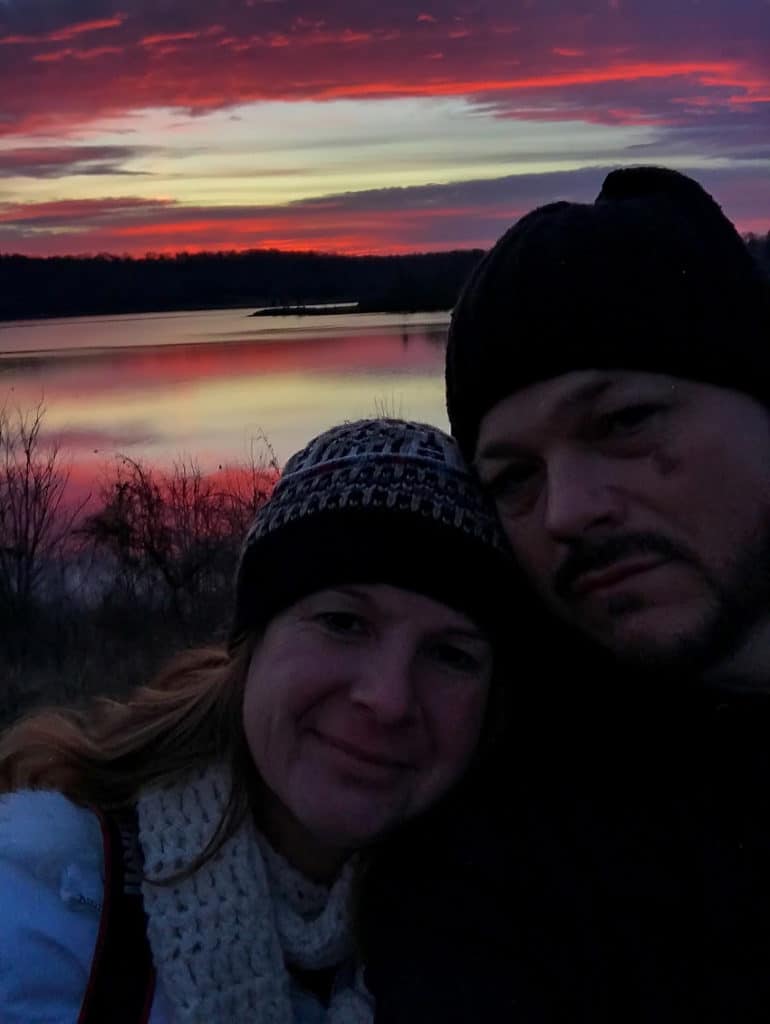 a couple standing in front of a pink and purple sunrise over a lake