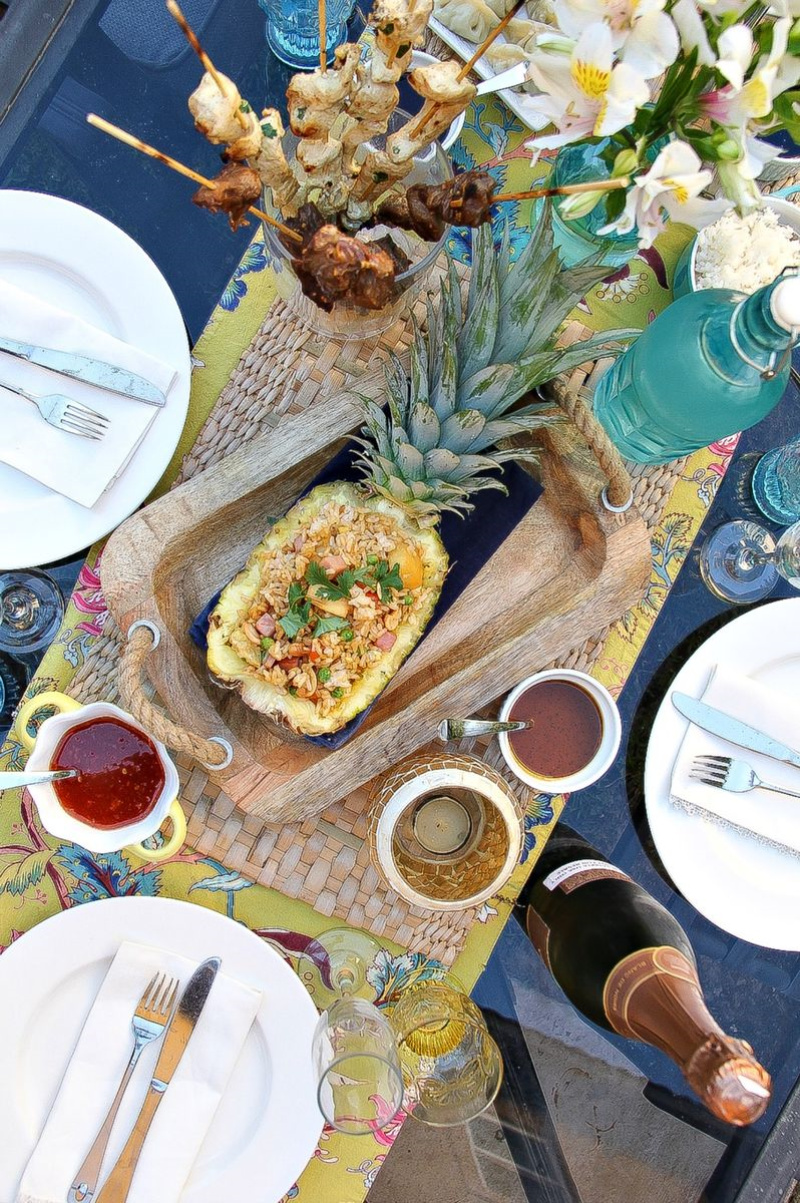 Looking down over an outdoor table with a half pineapple filled with pineapple fried rice, kabobs in a glass dish, flowers, and dining ware.