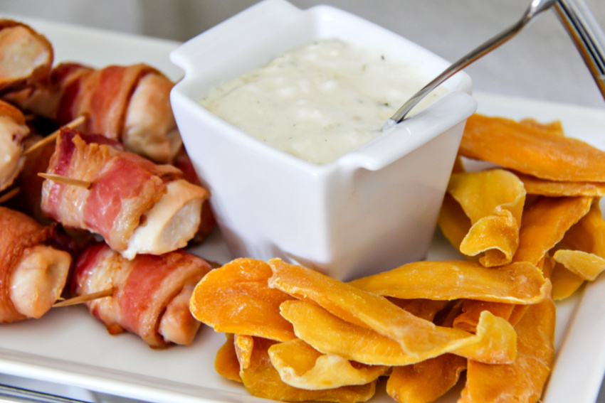 bacon wrapped chicken pieces with a dipping sauce in a white dish and dried mango