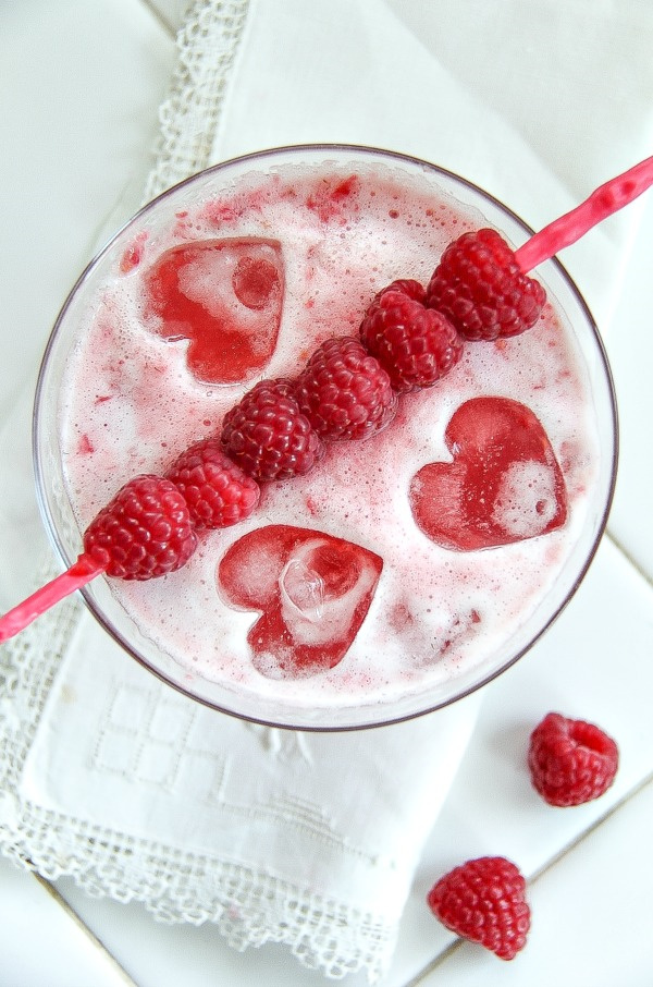a red cocktail garnished with raspberries and heart shaped ice cubes