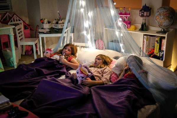 three girls on a mattress with purple blankets and a lighted canopy over the top for a sleepover