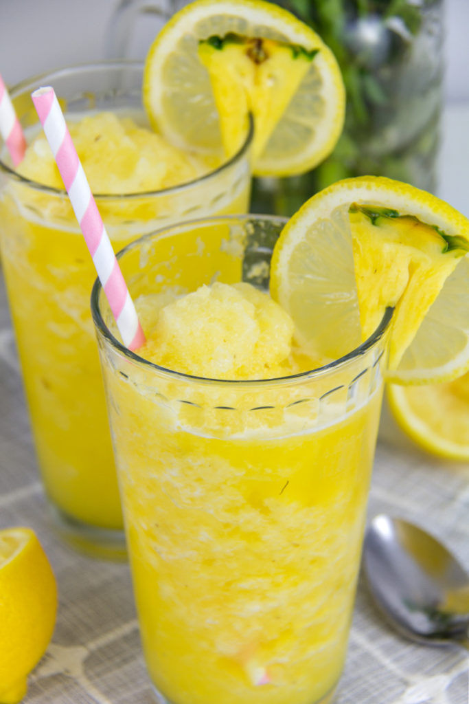 a pineapple and lemonade slushie summer drink for kids in a glass with a pink and white straw and garnished with lemon and pineapple
