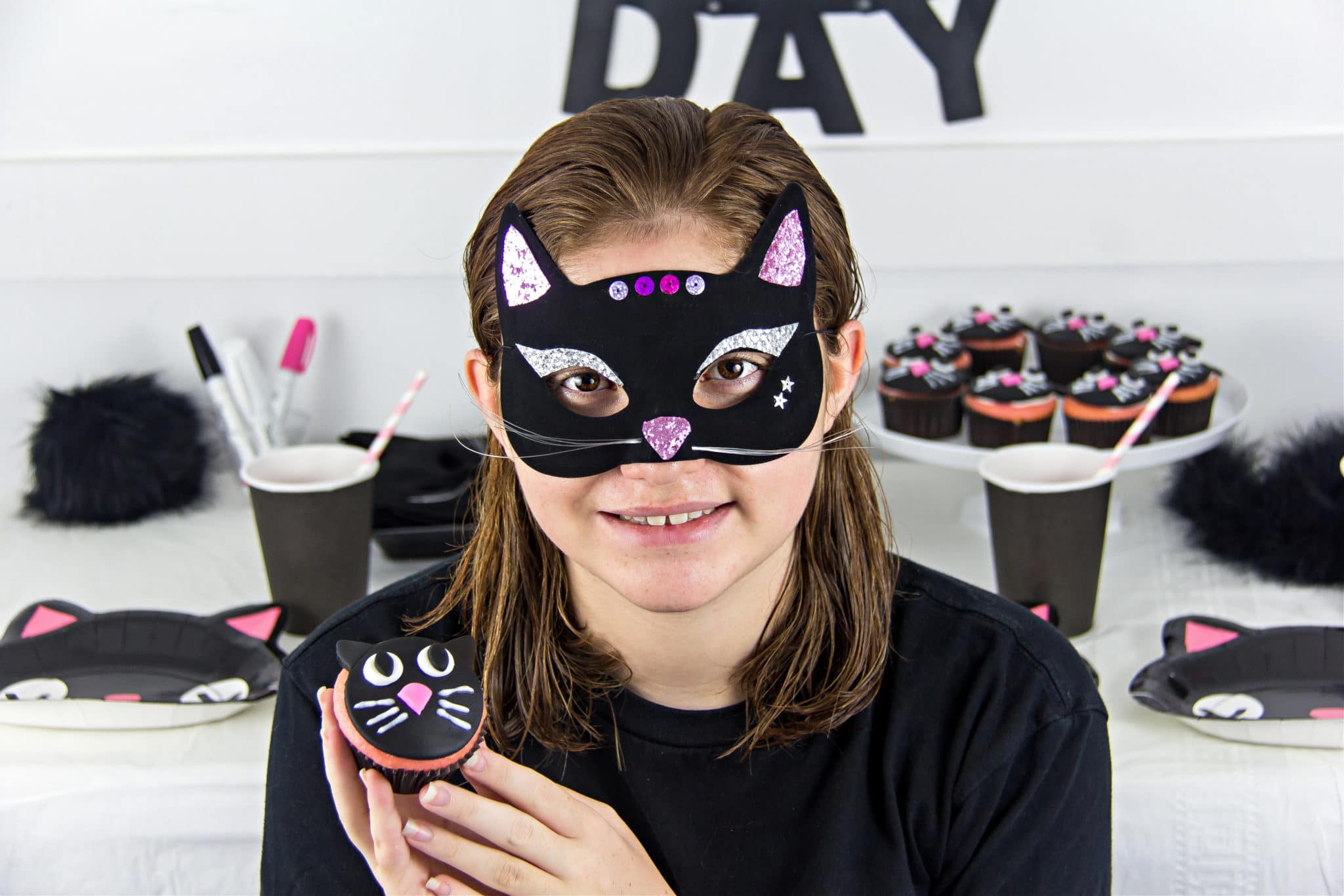 a girl wearing a homemade black cat face mask and holding a black cat cupcake for halloween