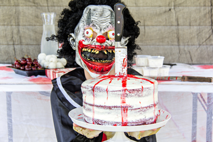 A boy wearing a slasher costume and holding a red velvet naked cake with a knife and blood on it.