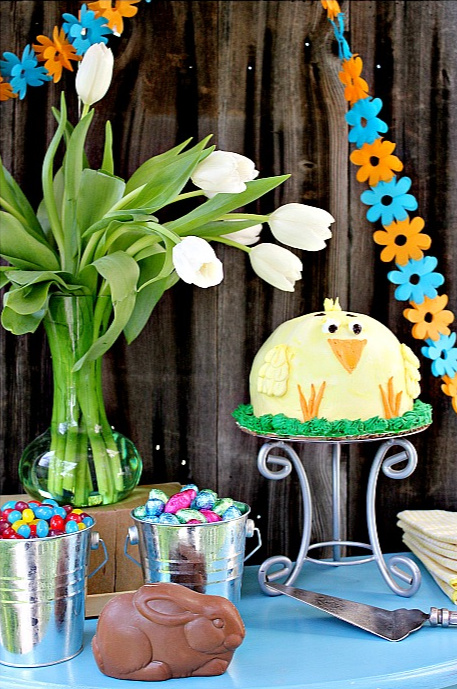 A blue table with a chocolate bunny, buckets of candy, and a baby chick ice cream cake for a party.