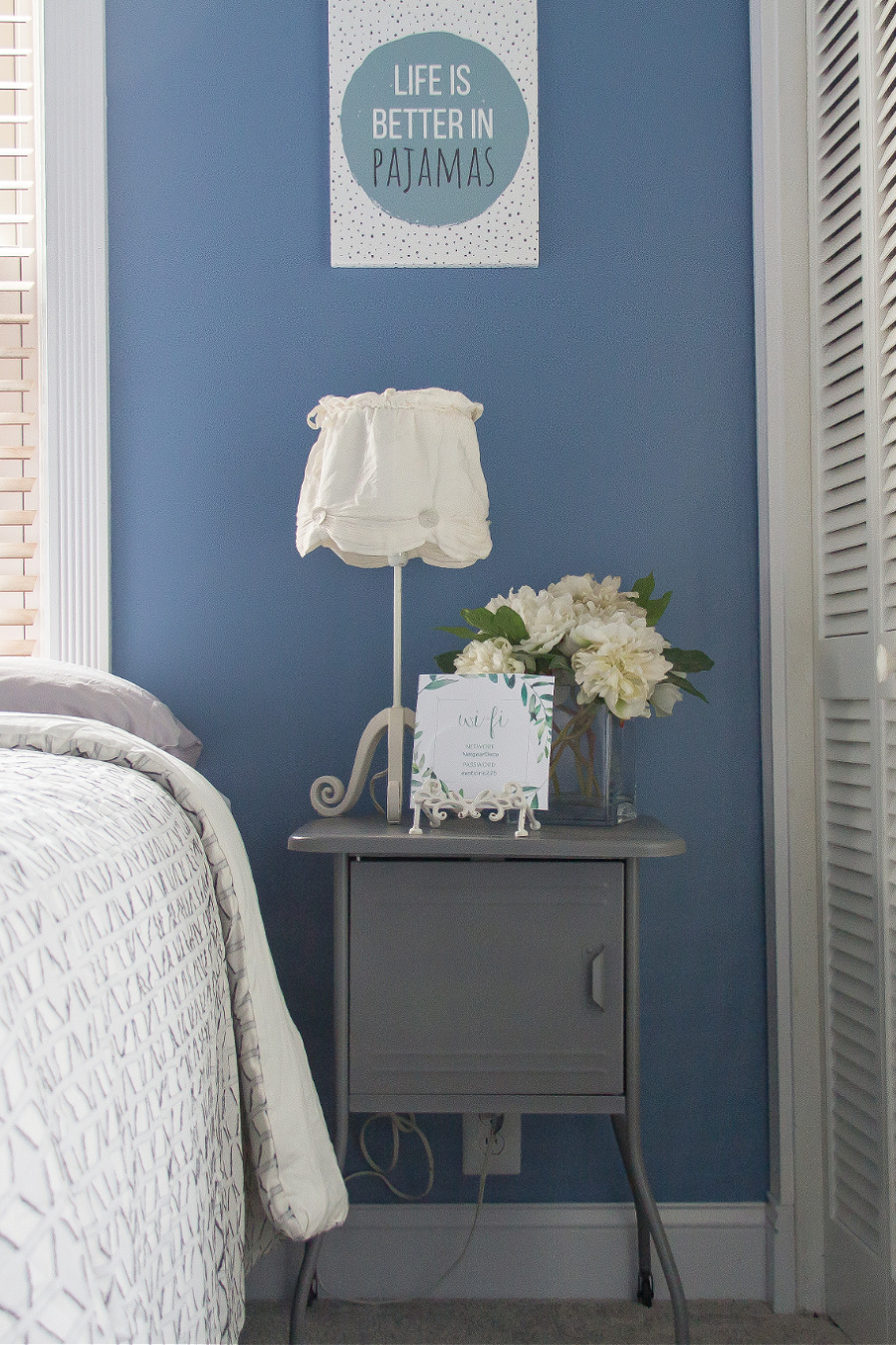 A grey metal bedside table with white flowers in a vase, a white bedside lamp, and a card with passwords for a guest bedroom.