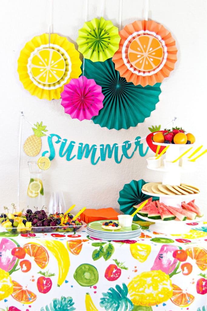 A party table for summer that is fruit themed with a tablecloth, decorations on the wall and fresh fruit ideas.