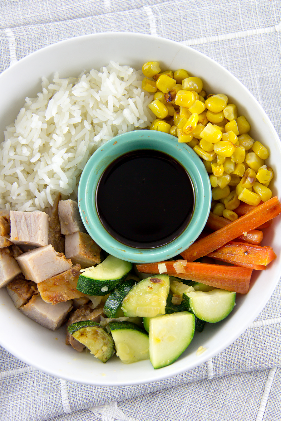 A bowl filled with rice, pork, zucchini, carrots and corn.