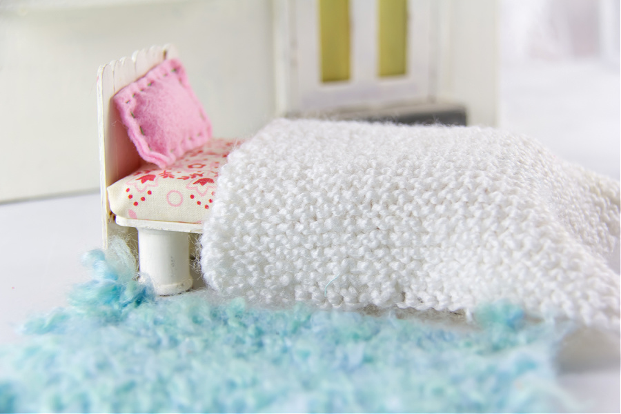 Handmade dollhouse bed with a hand knit blanket and floor rug.