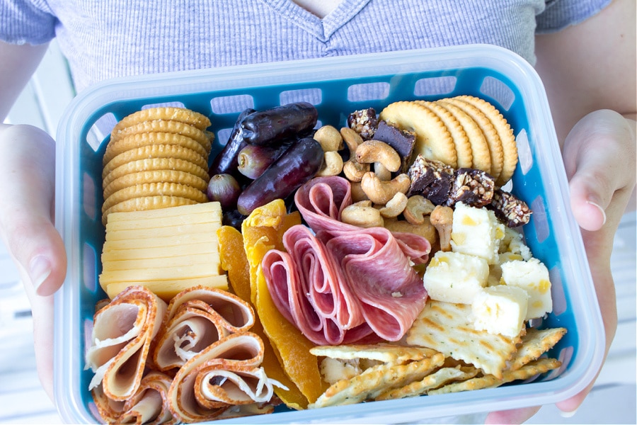 A charcuterie inspired lunch box for teenagers including meats, cheese, fruit, and nuts.