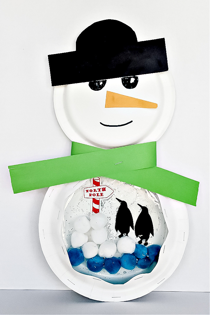 A paper plate snowman craft with pom poms and penguins in a paper plate snow globe.