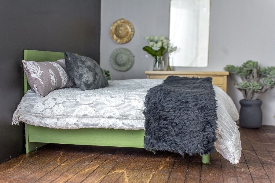 a green plywood bed with grey, black, and white bedding for a dollhouse.