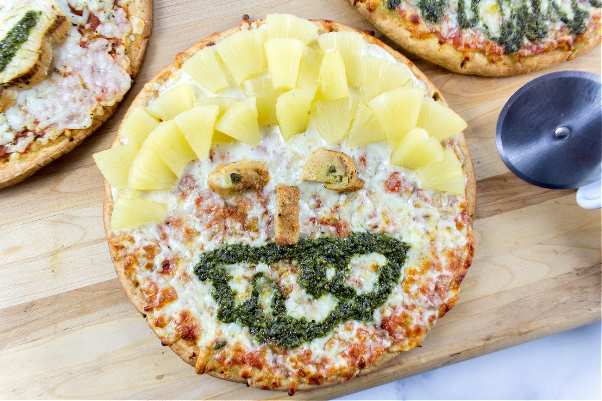 a funny face pizza decorated with pineapple for the hair, chicken nose and eyes, and pesto mouth.
