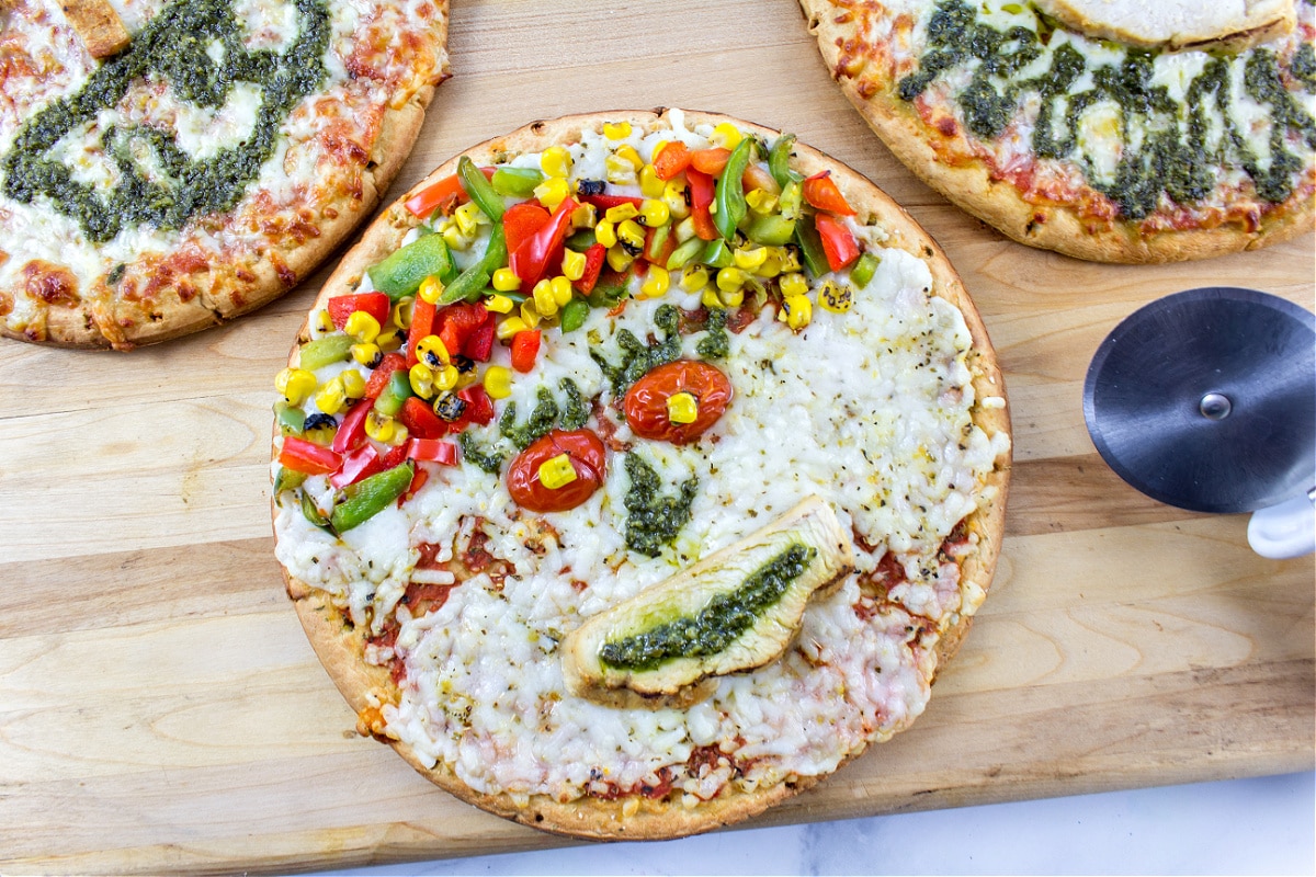 pizza decorating a funny face with mixed vegetables for the hair and chicken, tomato, and pesto for the eyes, nose, and mouth.