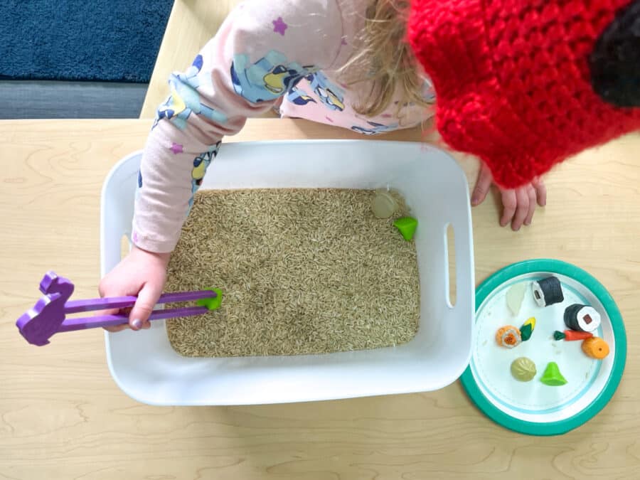 a small child using chopsticks to pick up rice and pretend food in a sensory tub