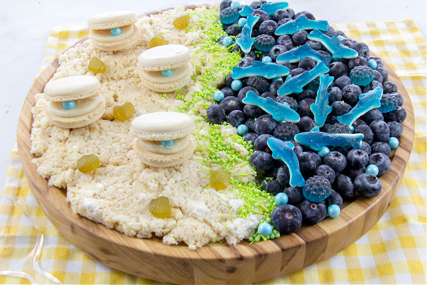 an ocean themed grazing board with gummy sharks, fruit, rice Krispies, and macaron clamshells