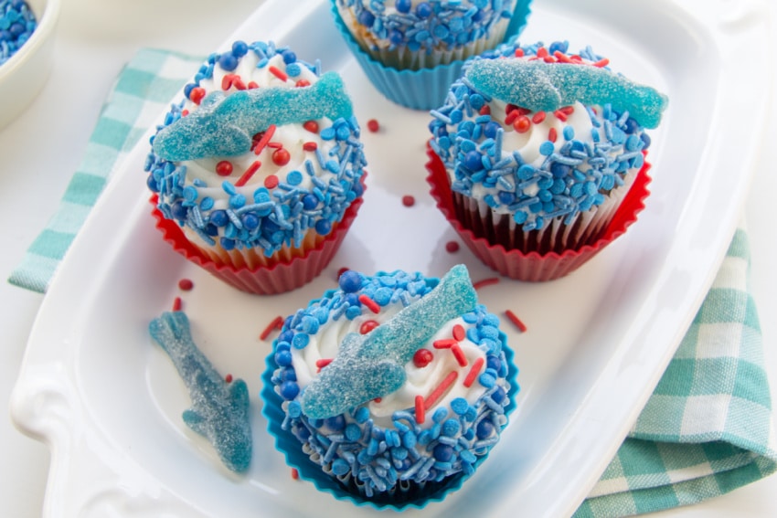 cupcakes decorated with red and blue sprinkles and topped with gummy shark for shark week